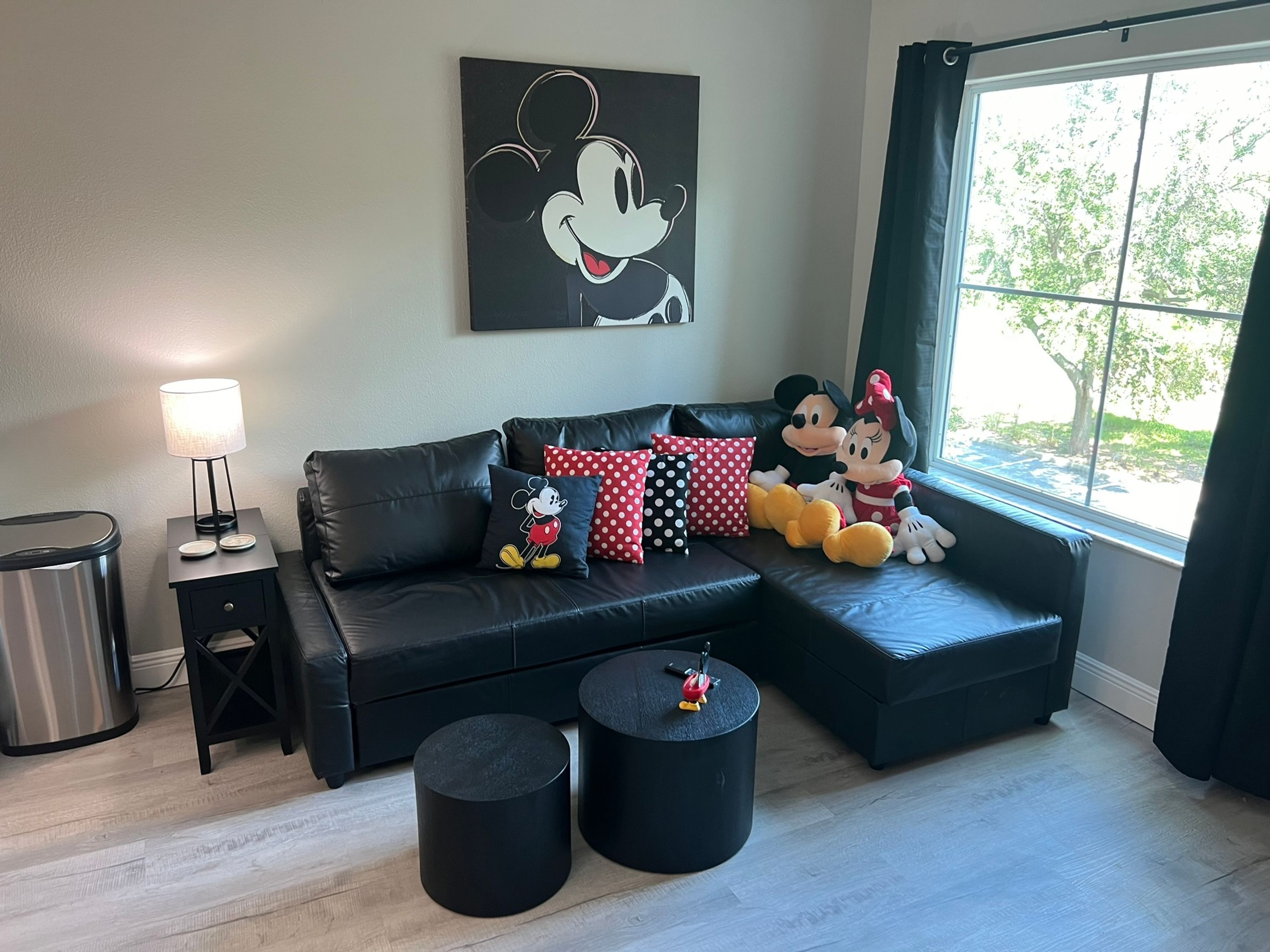 A modern living room with a black leather sofa, Mickey and Minnie Mouse plush toys, a lamp, and a large window with a view of trees.