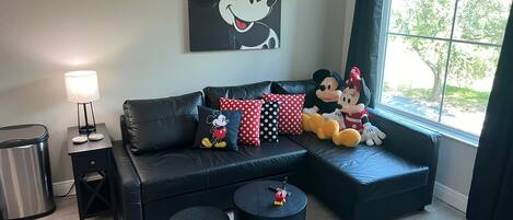 A modern living room with a black leather sofa, Mickey and Minnie Mouse plush toys, a lamp, and a large window with a view of trees.