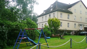A playground with a slide and swings, surrounded by a grassy area and a building with multiple windows. - Ferienwohnung Kemenate (Trendelburg OT Wülmersen)