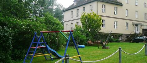 A playground with a slide and swings, surrounded by a grassy area and a building with multiple windows.