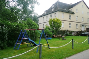 Ein Spielplatz mit Rutsche und Schaukeln, umgeben von einer Rasenfläche und einem Gebäude mit zahlreichen Fenstern.