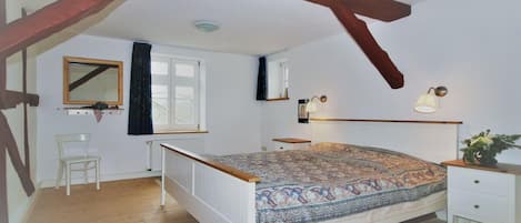 A bedroom with a wooden ceiling, a large bed with a patterned bedspread, a bedside table with a lamp, a chair, and a window with curtains.