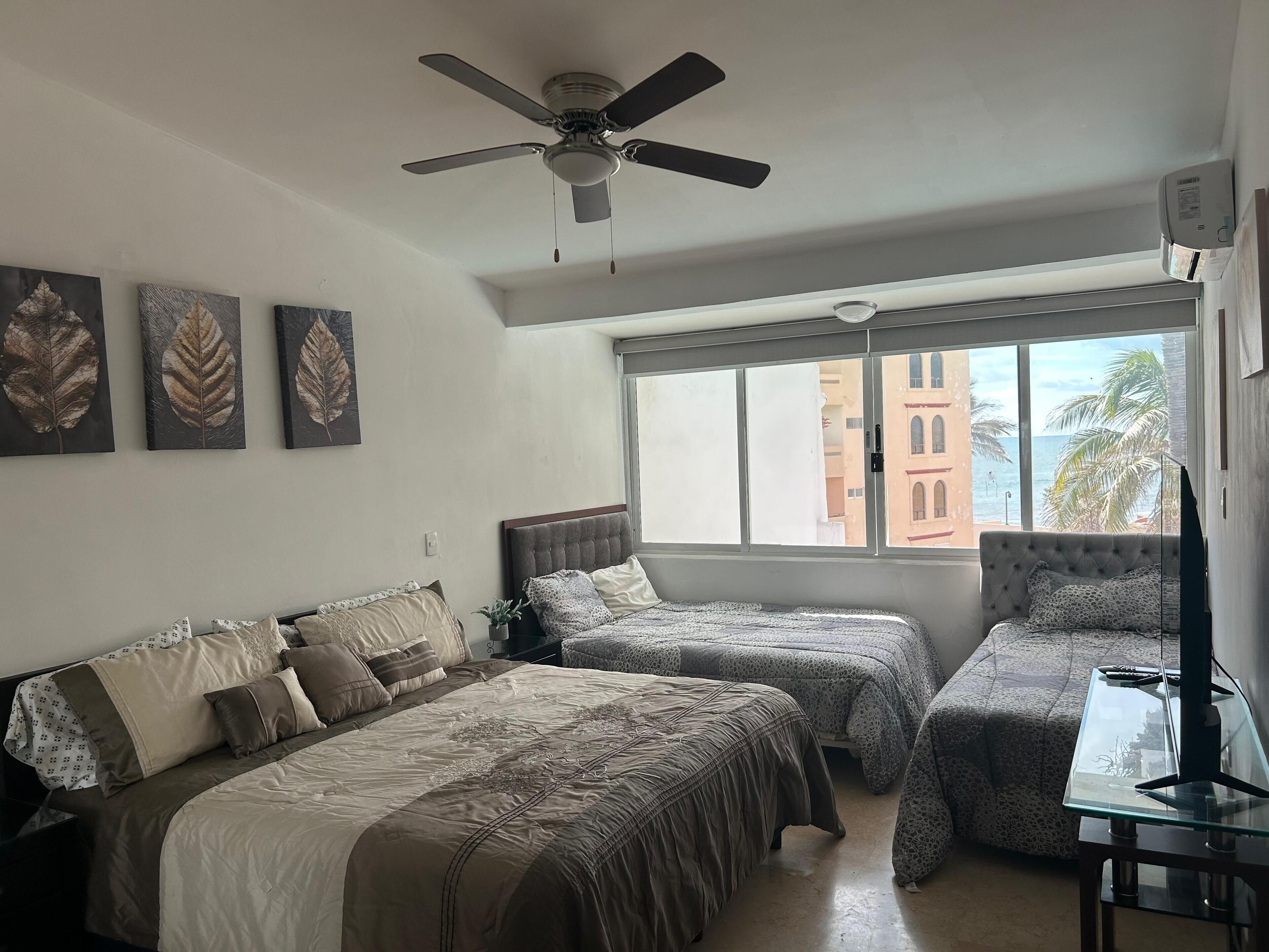 A bedroom with two beds, a ceiling fan, and a large window with a view of buildings and palm trees.