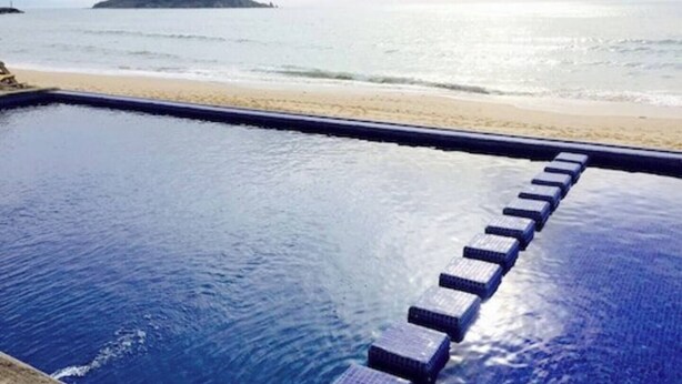 A pool with steps leading to the beach, a sandy shore, and a distant island. - Ocean front resort 2-bedroom condo in charming Mazatlán with AC, WiFi (Mazatlán)
