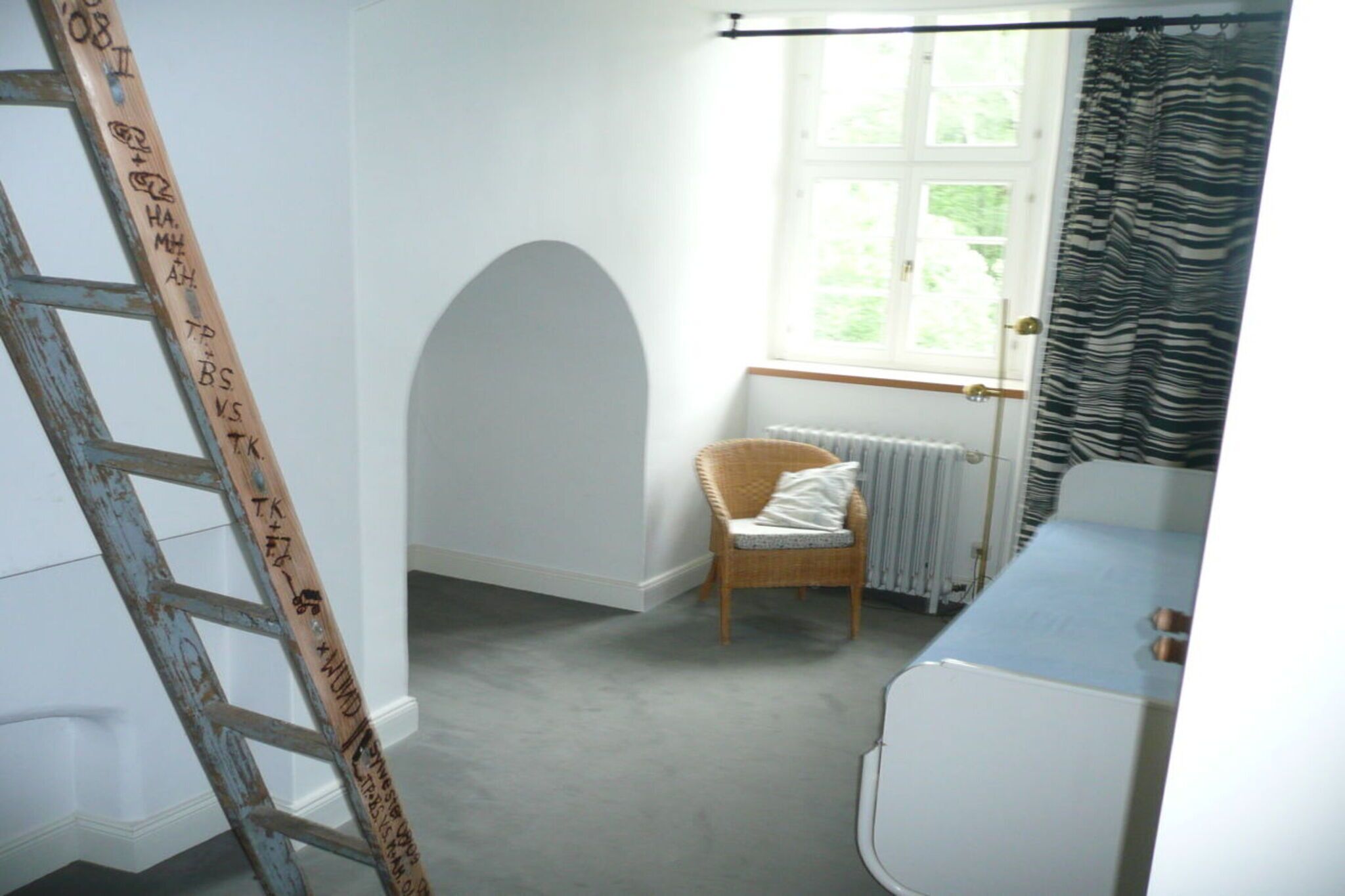 A room with a white wall, a wooden ladder, a wicker chair, a radiator, and a window with curtains.
