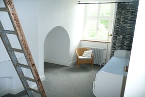 Una habitación con pared blanca, escalera de madera, silla de mimbre, radiador y ventana con cortinas.