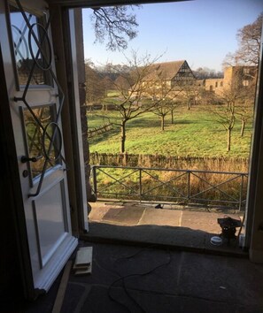Vista desde una habitación con puerta que da a un espacio al aire libre. La habitación tiene ventana con reja metálica y puerta con marco blanco. Se ve un campo cubierto de césped, árboles y un edificio a lo lejos.