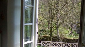 Blick aus einem Zimmer mit einem großen Fenster, einem Korbstuhl und einem weißen Metallgitter, das zu einem Garten führt.