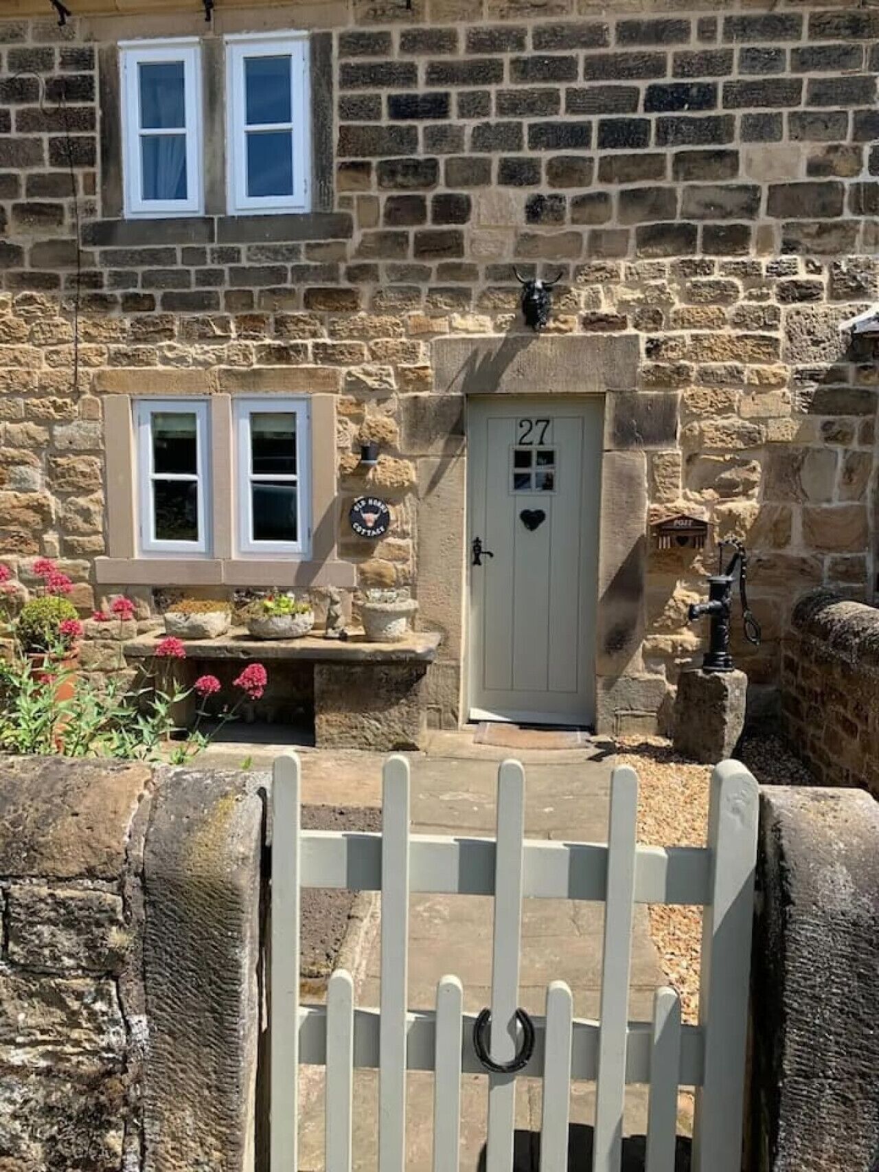 A stone house with a white door marked with the number 27, a small porch with potted plants, and a white gate.