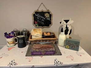 A table with a welcome sign, a basket of bread, a mug labeled 'cream', a milk jug, and a card with a cow illustration.