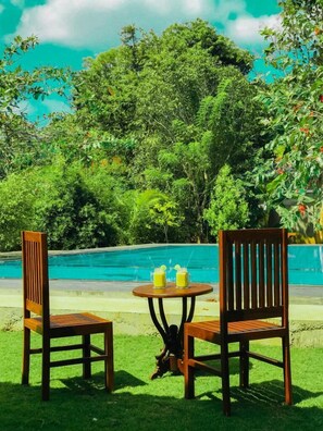 Two wooden chairs and a small round table with two glasses of yellow beverage by a swimming pool surrounded by lush greenery. - NATURALIZA (Dambulla)