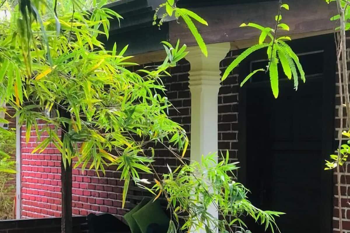 Un edificio de ladrillo con un porche cubierto, un banco verde y un pequeño jardín con plantas y flores.