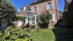 Ein zweistöckiges Steinhaus mit Terrasse, einer Glastür, die zu einem überdachten Sitzbereich im Freien führt, und einem gepflegten Garten.