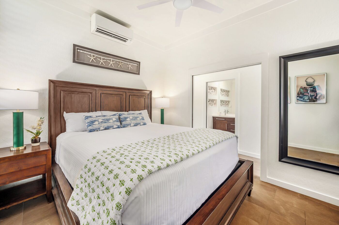 Une chambre d’hôtel avec un lit en bois, une table de chevet avec une lampe, un grand miroir et un climatiseur fixé au mur.