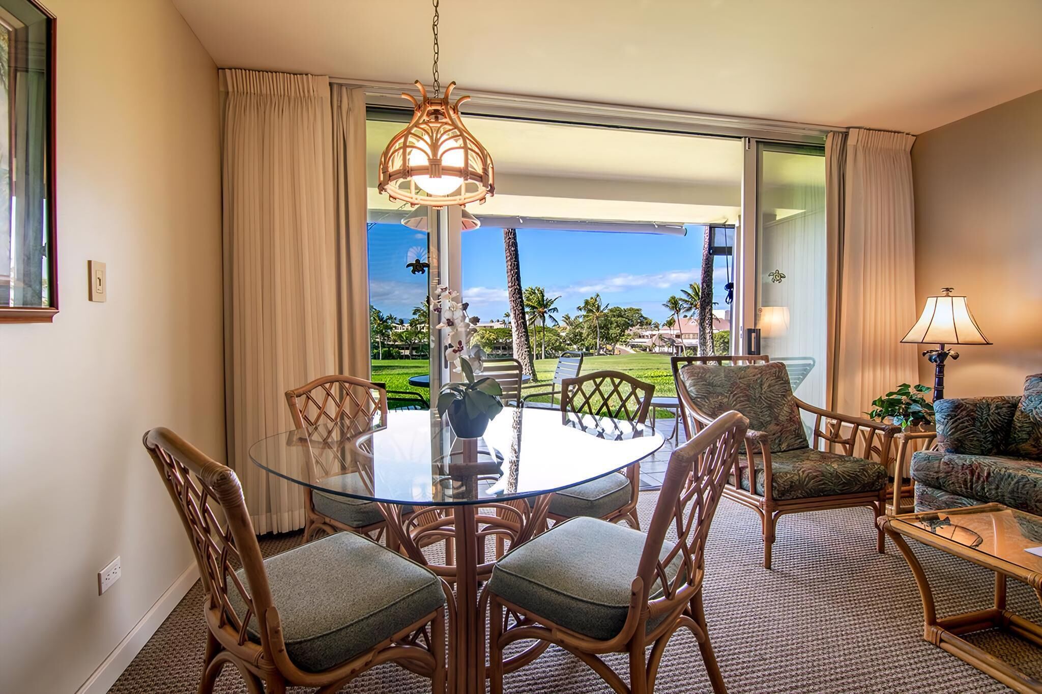 A hotel room with a glass dining table surrounded by wicker chairs, a sofa, and a lamp.