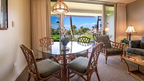 Habitación de hotel con una mesa de comedor de vidrio rodeada de sillas de mimbre, un sofá y una lámpara.