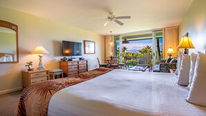 Habitación de hotel con una cama grande, ventilador de techo, televisor, escritorio con lámpara y vista al exterior.