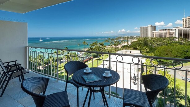 A balcony with a view of the ocean and cityscape, furnished with wicker chairs and a table. - Waikiki Shore 1016 Ocean View 1 Bedroom (Honolulu)