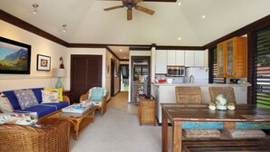 A living room with a sofa, coffee table, and dining table. There is a kitchen area with cabinets and a microwave.