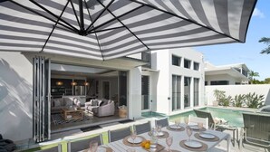 An outdoor dining area with tables set for a meal, a pool, and a modern house in the background. - Frangipani Beach House (Kingscliff)
