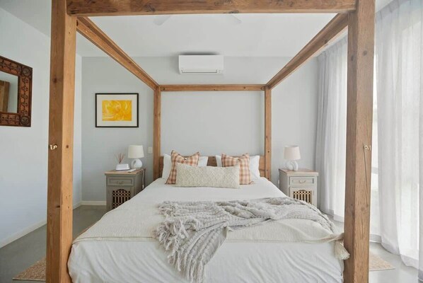 A bedroom with a four-poster bed, two bedside tables, a nightstand, a wall-mounted air conditioner, and a framed picture on the wall.