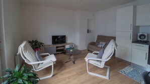 A living room with two white chairs, a glass coffee table, a beige sofa, a TV, a plant, and a kitchen area with cabinets and a microwave.