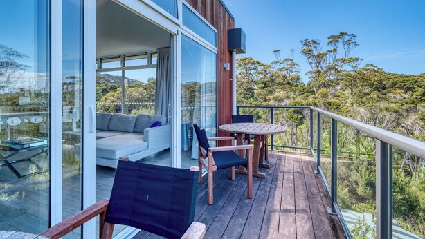 A modern outdoor deck with a glass door leading to a living area, a round wooden table, and chairs. - The Peninsula Grande (Karikari Peninsula)
