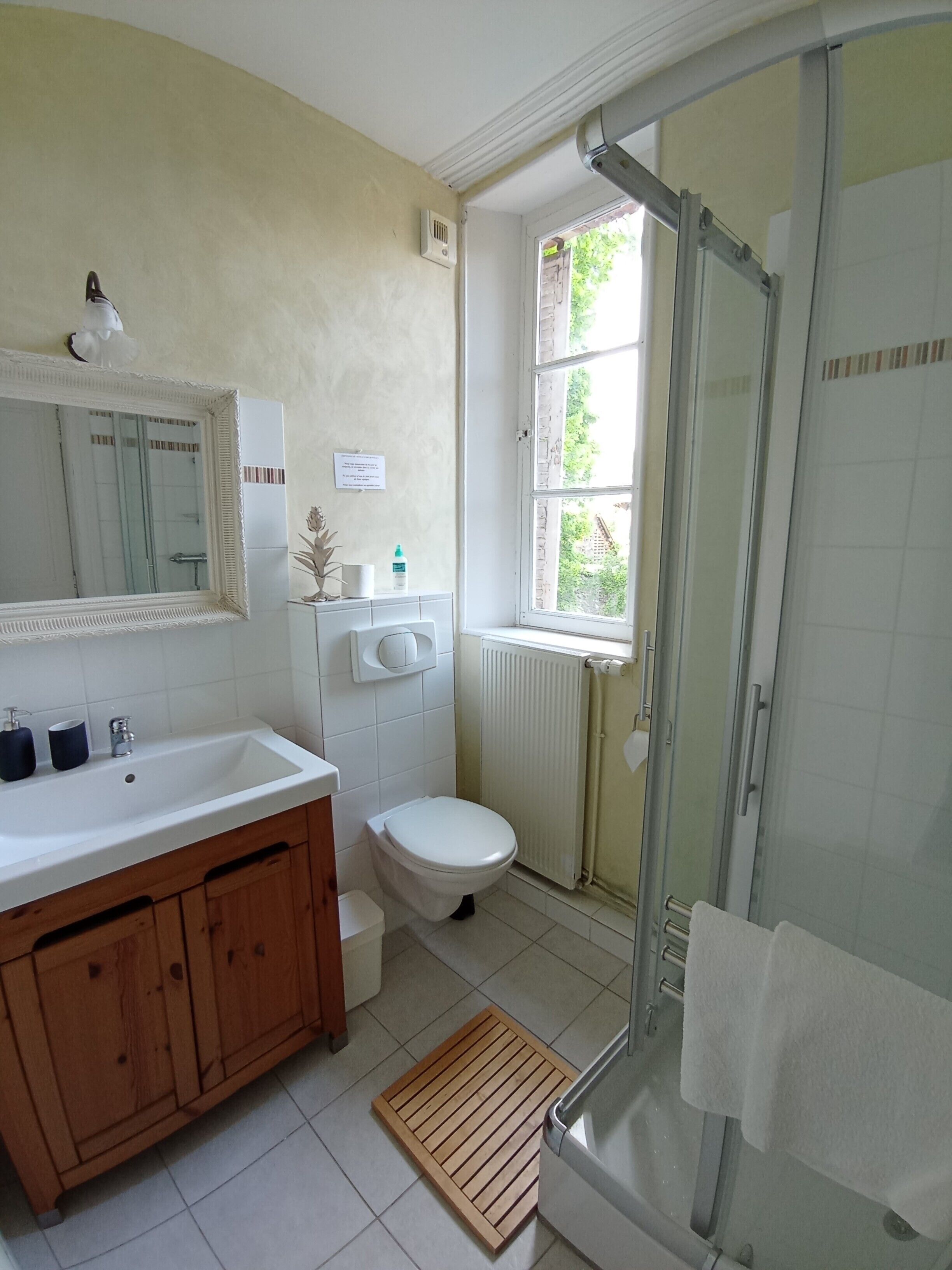 A bathroom with a wooden vanity, a white sink, a toilet, a glass shower enclosure, and a window with a view of greenery outside.