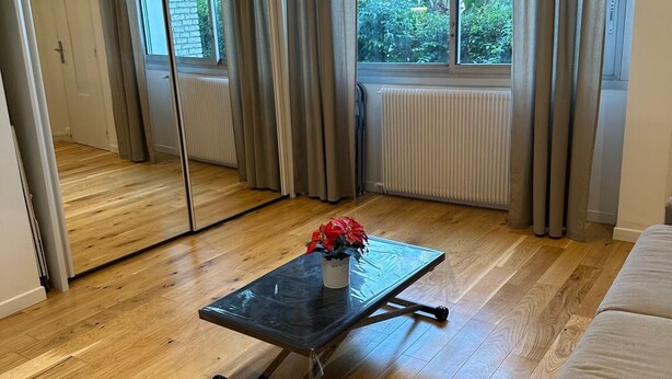 A room with a wooden floor, a glass-top table with a red flower vase, a grey sofa, and a large window with curtains. - The blue tide  (Nice)
