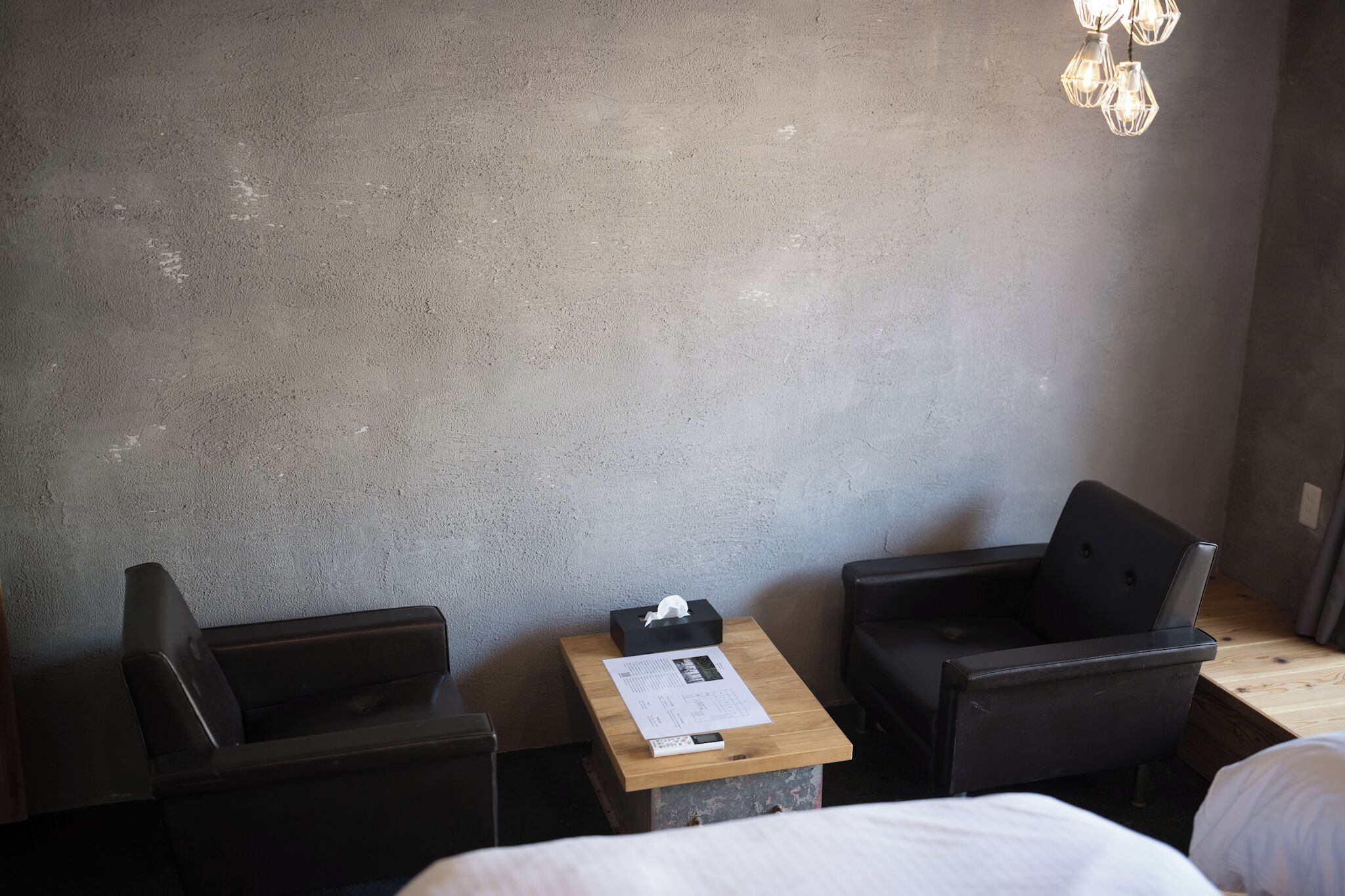 A hotel room with a bed, two black armchairs, a small wooden table with a tissue box and a receipt, and a wall-mounted light fixture.
