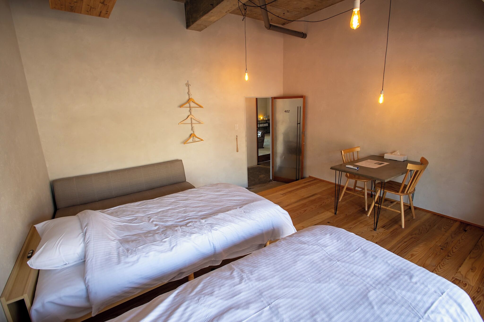 A room with two beds, a wooden dining table with chairs, a refrigerator, and a hanging light fixture.