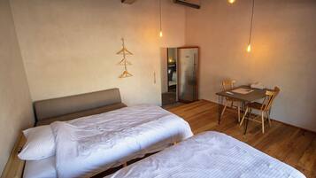 A room with two beds, a wooden dining table with chairs, a refrigerator, and a hanging light fixture.