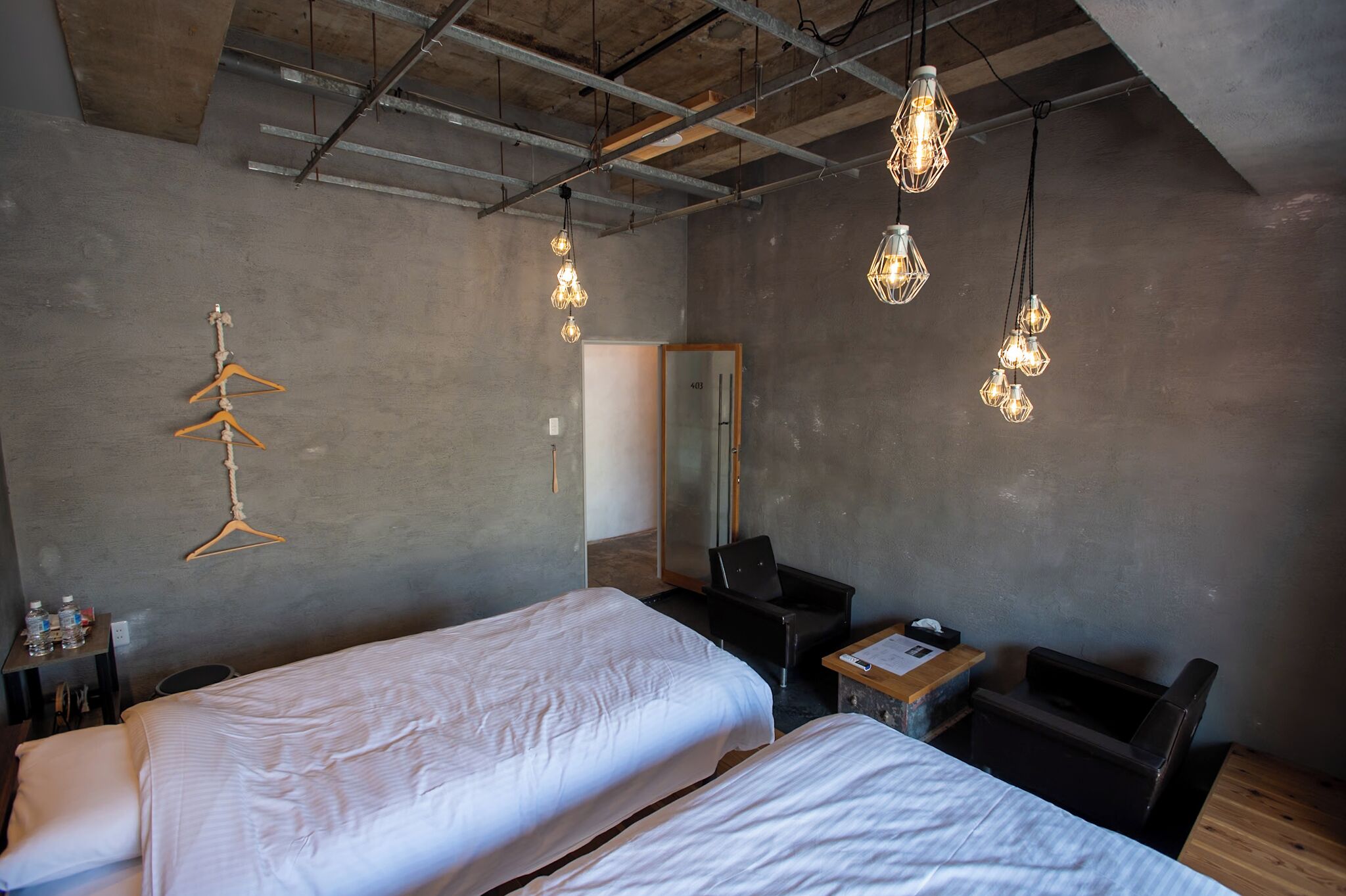 A hotel room with two beds, a desk, a chair, a hanging light fixture, and a coat rack.