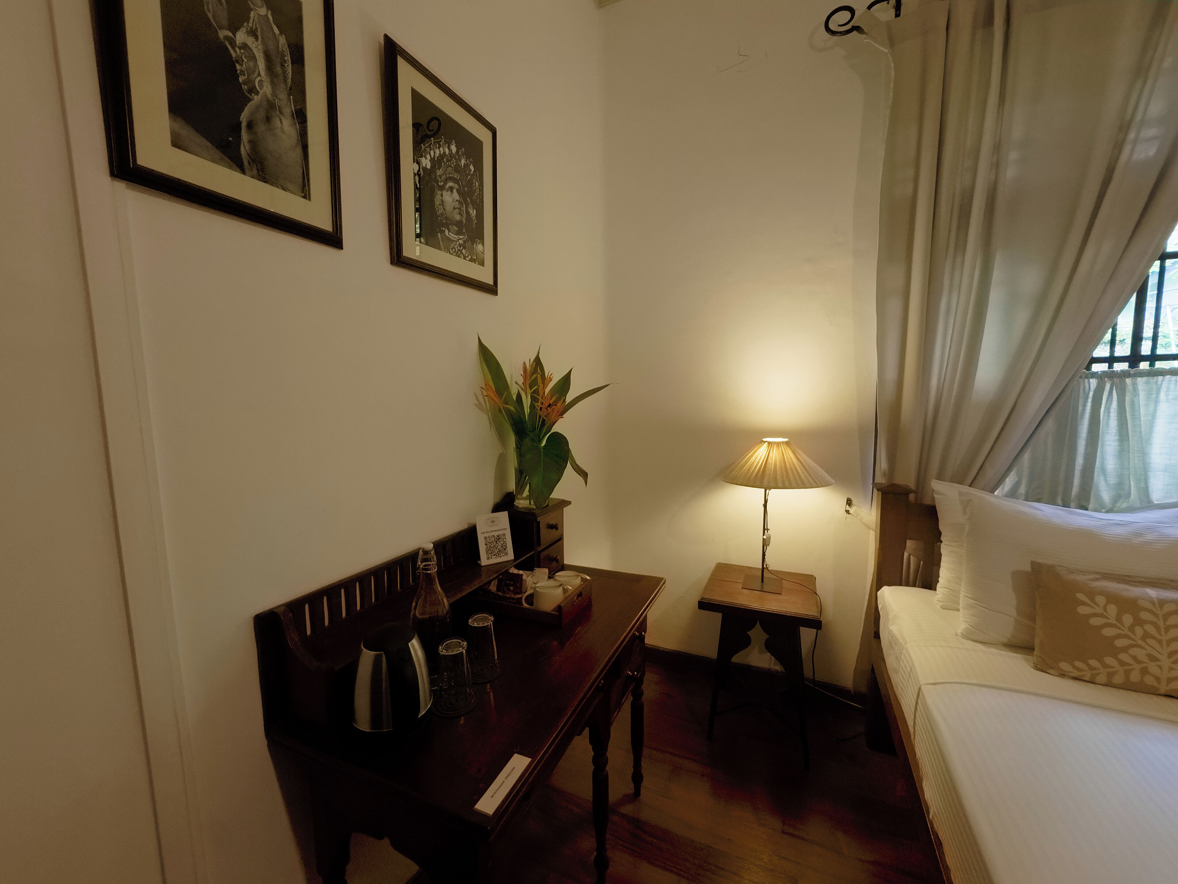 A hotel room with a bed, a desk with a lamp, a small table, and two framed pictures on the wall.