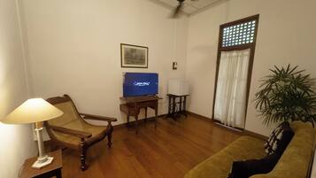 A living room with a wooden floor, a sofa, a chair, a table with a lamp, a television, a ceiling fan, and a window with a curtain.