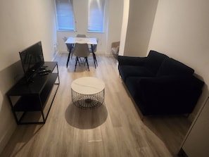 Een moderne woonkamer met een televisie, een ronde salontafel, een zwarte bank, een eettafel met stoelen en een raam met jaloezieën.