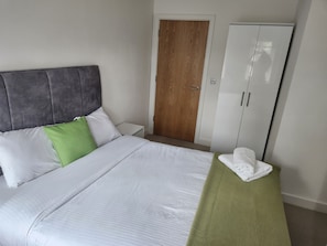 Een net opgemaakt bed met witte en groene kussens, een grijze hoofdbord en een groene handdoek op een klein tafeltje ernaast.