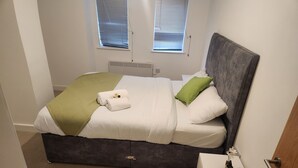 A neatly made bed with a green throw, white pillows, and a grey headboard in a room with a white door and a small white table with a plant. - Modern & Cozy 1bed Apartment Close to City Centre (Coventry)