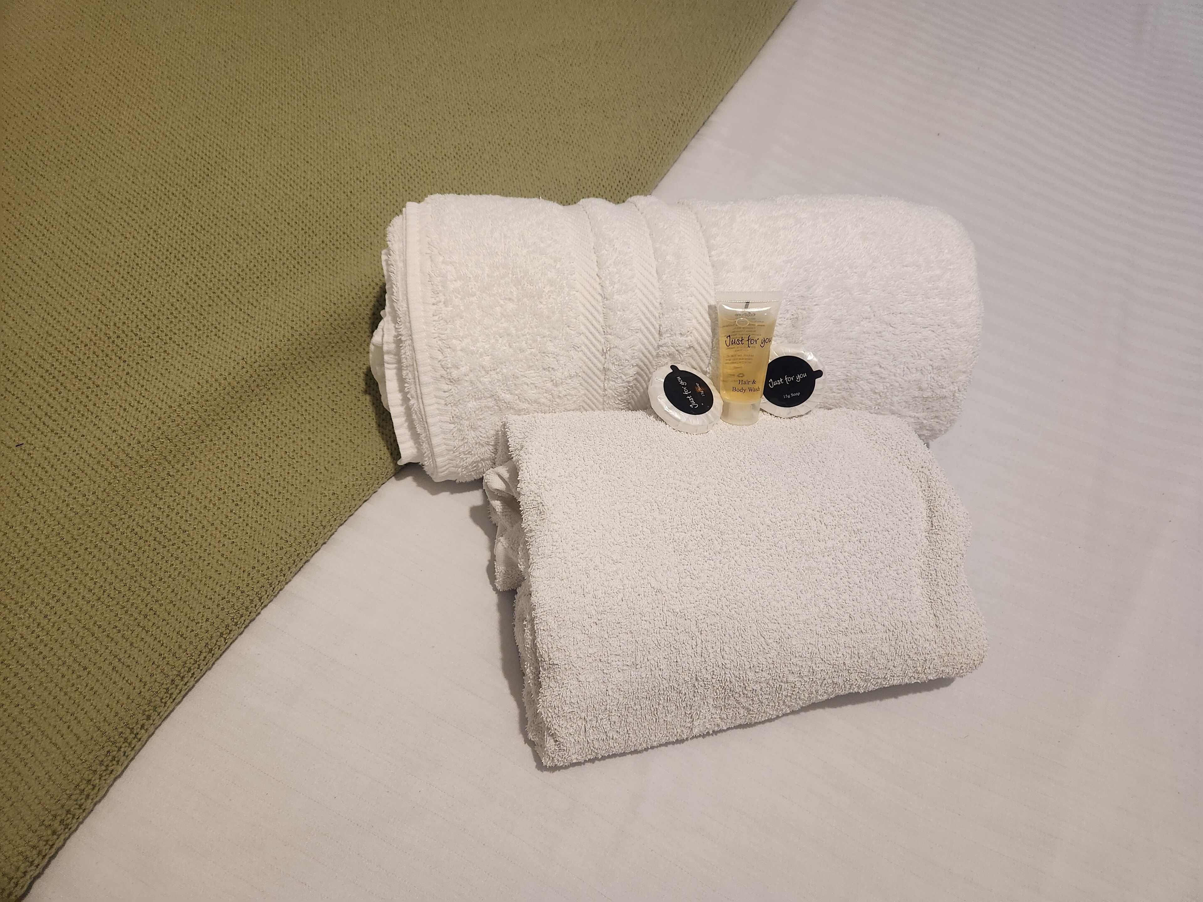 Une serviette blanche, soigneusement pliée, avec une petite bouteille de gel pour les cheveux et le corps posée dessus, est placée sur un lit agrémenté d’un oreiller vert à texture et d’un oreiller blanc.