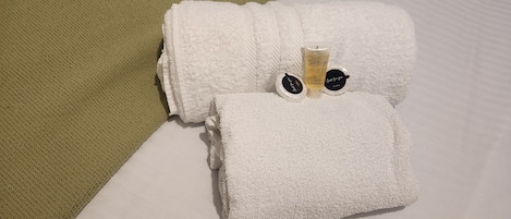 Une serviette blanche, soigneusement pliée, avec une petite bouteille de gel pour les cheveux et le corps posée dessus, est placée sur un lit agrémenté d’un oreiller vert à texture et d’un oreiller blanc.