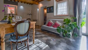 A cozy room with a wooden dining table, a sofa with pink cushions, and a large window with curtains. - Cosy apartment for 4 guests with private pool, A/C, WIFI, TV, patio and pets allowed (Capannori)