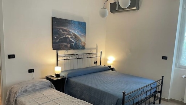 A bedroom with two beds, a nightstand with a lamp, a wall-mounted light fixture, and a painting of a celestial scene.