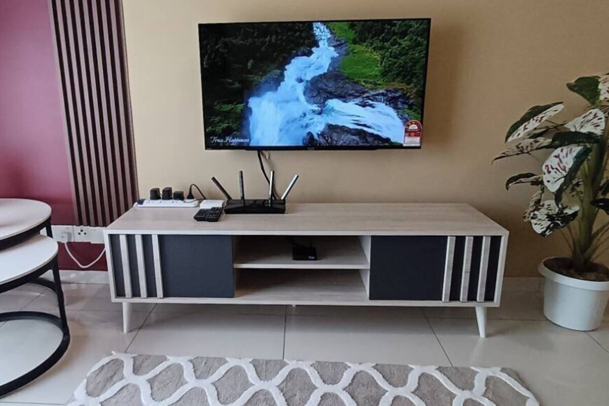 Een woonkamer met een aan de muur bevestigde televisie, een ronde tafel met twee witte stoelen, een witte bank, een potplant en een kamerbreed tapijt met een patroon.