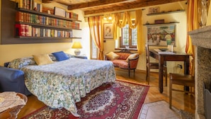 Ein gemütliches Zimmer mit Holzbalkendecke, einem Bett mit floralem Bettzeug, einem Sessel, einem Schreibtisch und einem Bücherregal voller Bücher.