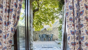 Blick durch ein Fenster mit blumigen Vorhängen auf eine Steinmauer und einen Tisch mit Stühlen draußen.