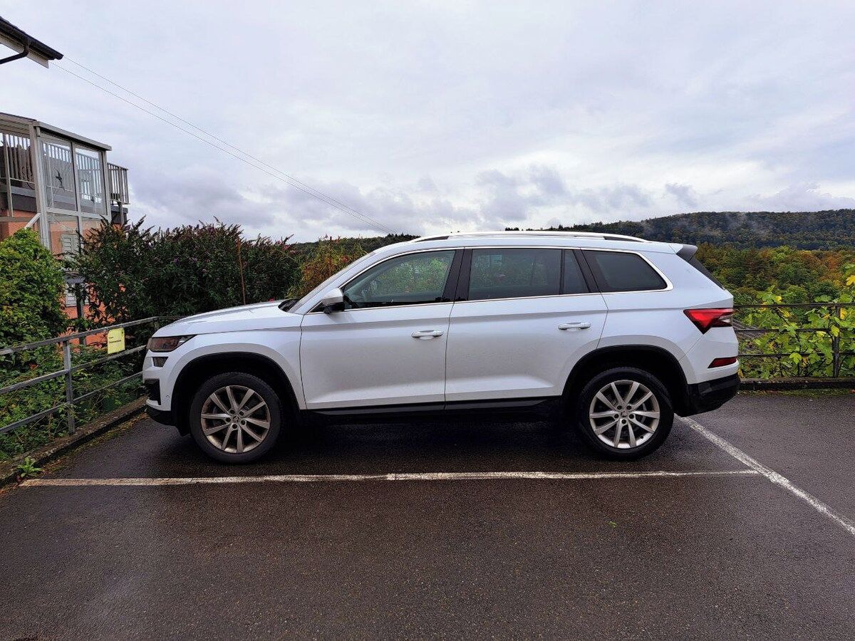 Ein weißer SUV steht auf einem Parkplatz, im Hintergrund sind ein Gebäude und Bäume zu sehen.