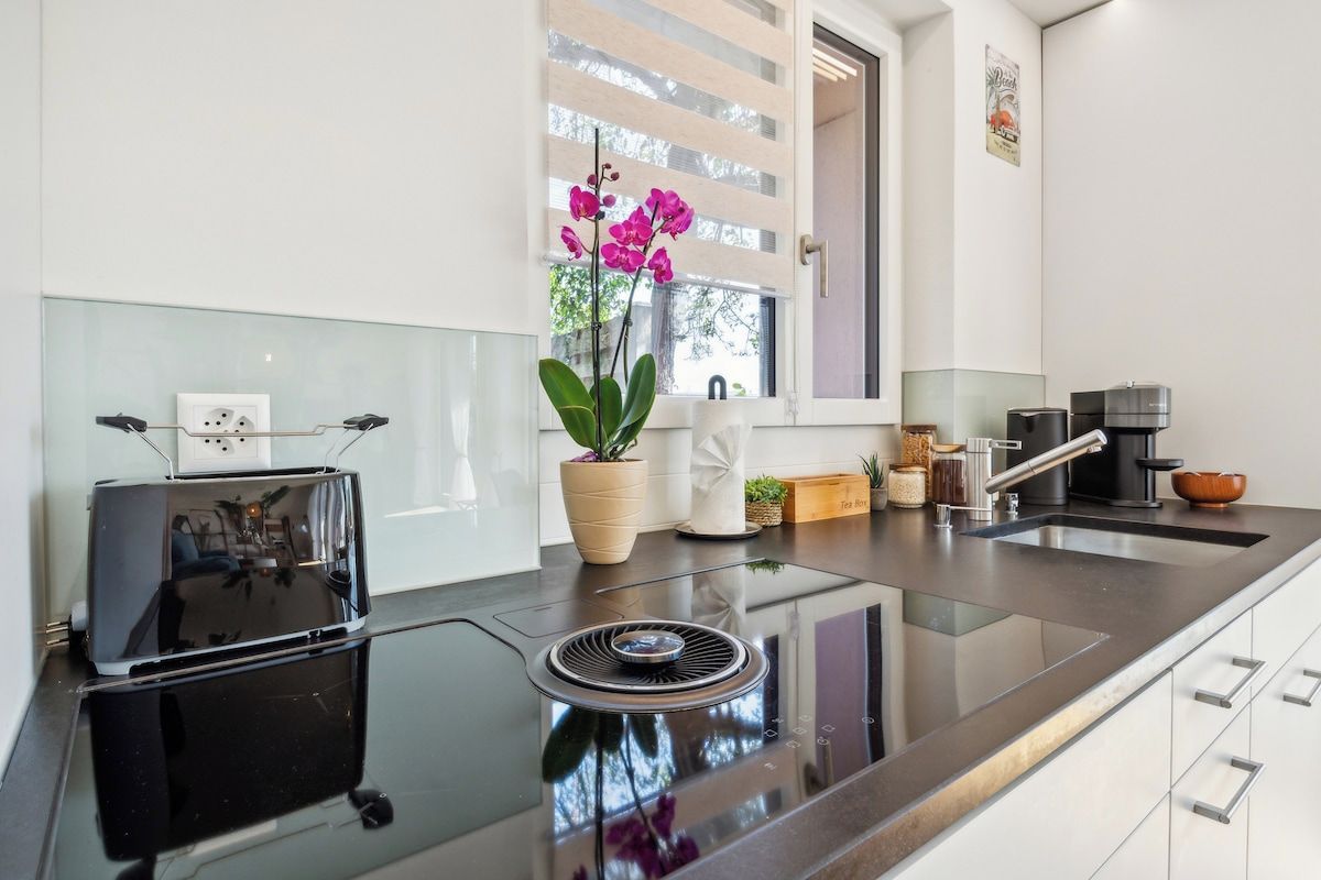 A modern kitchen with a black induction cooktop, a built-in oven, a window with blinds, and a potted orchid plant.