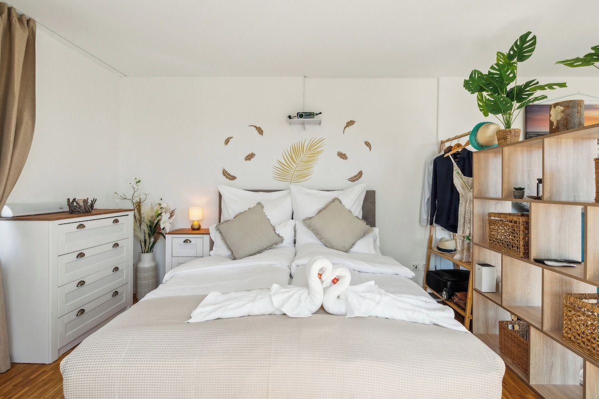 Una camera d'albergo con un letto, un comodino, una cassettiera, una mensola in legno contenitore e una pianta grande.