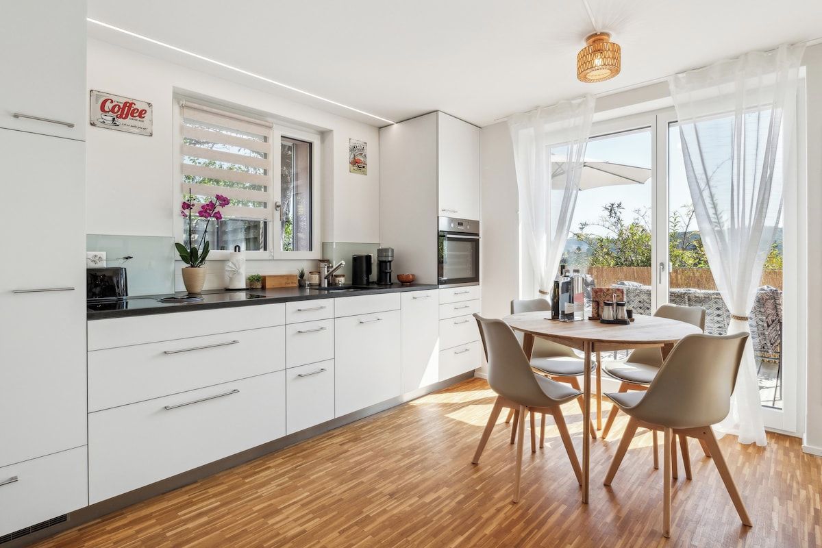 Eine moderne Küche mit weißen Schränken, ein Essbereich mit einem runden Tisch und Stühlen sowie ein Blick durch große Fenster.