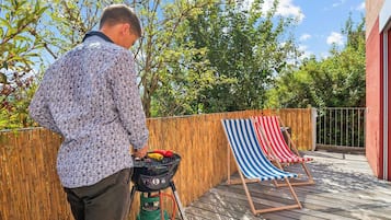 Jemand grillt draußen, mit einem Grill, einer Holzterrasse und einem gestreiften Liegestuhl.